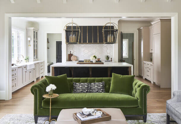 A Southern moern farmhouse kitchen with soft, off-white cabinets, a black metal hood, dark gray kitchen island, and olive green accents including the green couch in the neighboring living room.